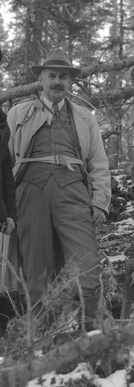 Black and white photograph of a man in a suit, with a hat and a brush moustache, posing in a forest.