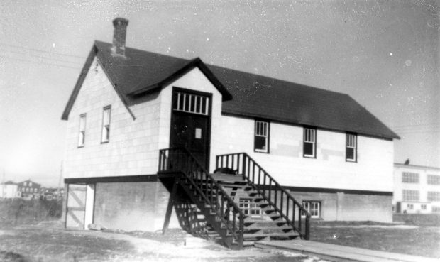 Black and white photograph of a two-storey white church. A staircase, on which a dog is asleep, leads to the main door. On the side, an open door gives access to the basement. In the background on the right, the Catholic primary school.