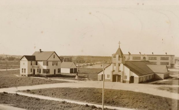 Sepia photograph of the presbytery and the church. The church is low in height and a large cross overhangs the main entrance. In the background, the Catholic primary school.