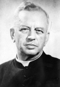 Black and white photograph of an elderly man in a black shirt and Roman collar.