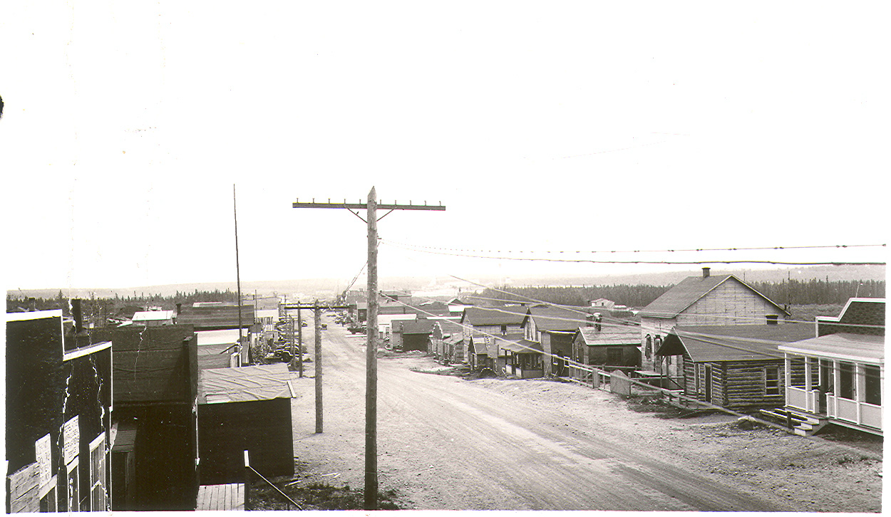 Probably taken from the roof of a building, a black and white photograph of a gravel road lined with wooden log and plank buildings. A forest is visible in the background.