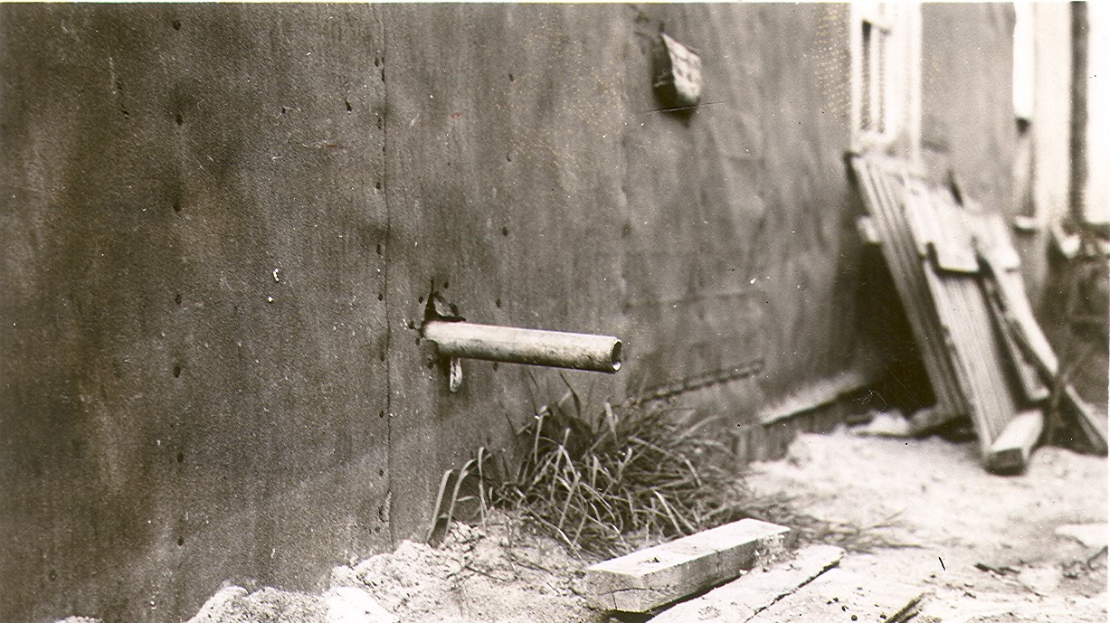Black and white photograph of a pipe coming out of a building covered with tar paper and spilling its contents onto wooden planks. In the background, a pile of wood.