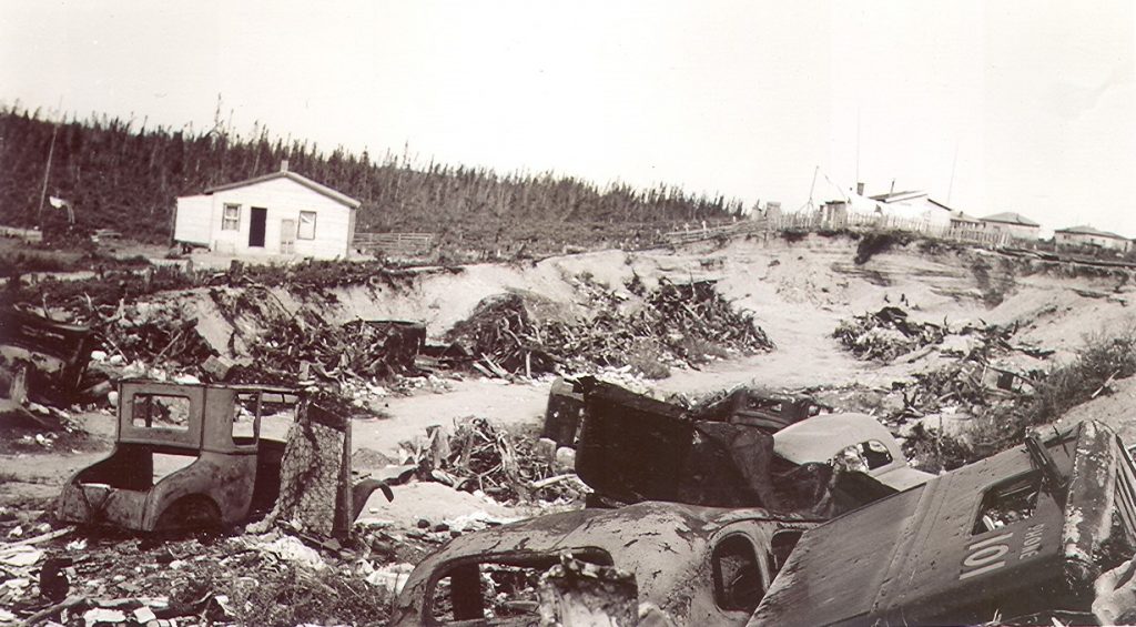 Photographie en noir et blanc d’une fosse de sable rempli de carcasses de voitures rouillées et de différents déchets. En arrière-plan, plusieurs résidences derrière une clôture. 