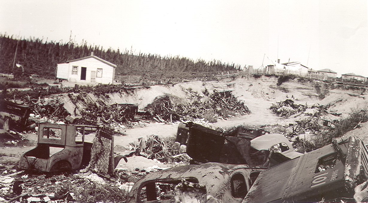 Black and white photograph of a sand pit filled with rusted car bodies and a variety of waste. In the background, several residences behind a fence.