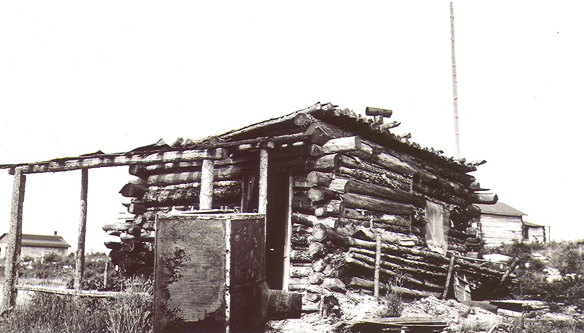 A black and white photograph of a rudimentary log cabin that is collapsing. In the background, other buildings that are similar, but in better condition.
