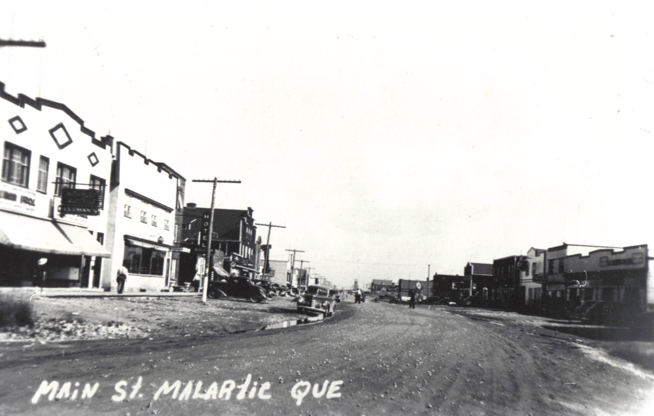Black and white photograph of a wide gravel road lined with good-quality buildings. On the left, the sign for Cleman’s store and a hotel. Several cars are parked on either side. In the background, the headframe of a mine shaft.