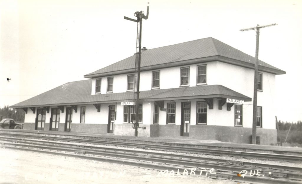 Black and white photograph of a good-quality, two-storey building. In two places, posters bearing the name of the locality: 