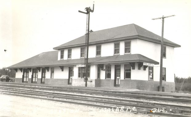 Black and white photograph of a good-quality, two-storey building. In two places, posters bearing the name of the locality: Malartic. In the foreground, several railway tracks. The photo bears the inscription Malartic Que.