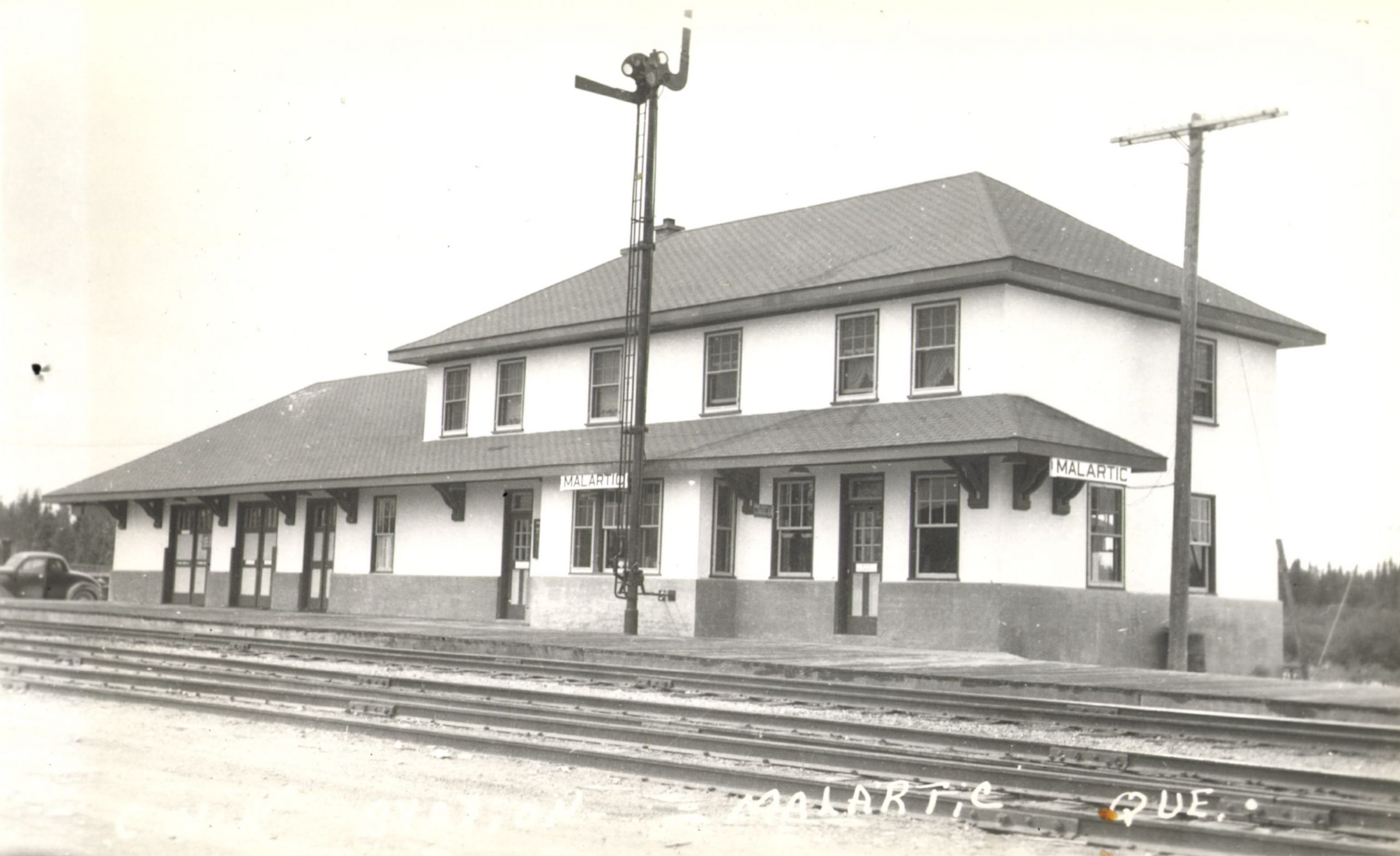 Black and white photograph of a good-quality, two-storey building. In two places, posters bearing the name of the locality: "Malartic". In the foreground, several railway tracks. The photo bears the inscription "Malartic Que".