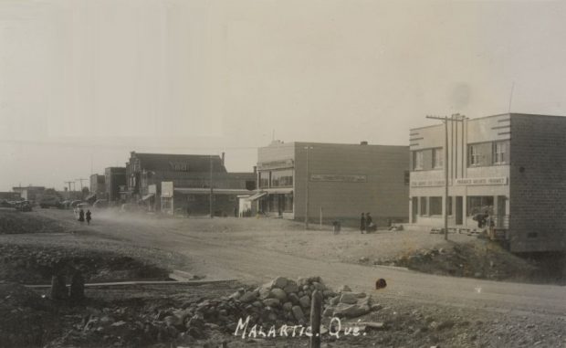 Black and white photograph of a gravel road under construction with a row of recently-constructed buildings in the background. Two women walking behind a car that raises a cloud of dust. In the foreground, a bridge and a small pile of rocks. The photograph bears the inscription Malartic, Qué.