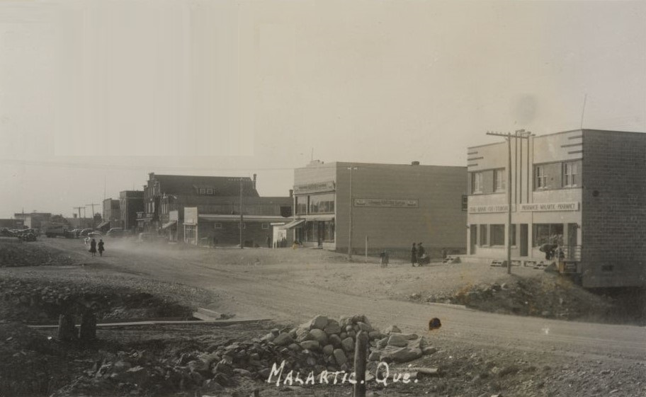 Black and white photograph of a gravel road under construction with a row of recently-constructed buildings in the background. Two women walking behind a car that raises a cloud of dust. In the foreground, a bridge and a small pile of rocks. The photograph bears the inscription "Malartic, Qué".