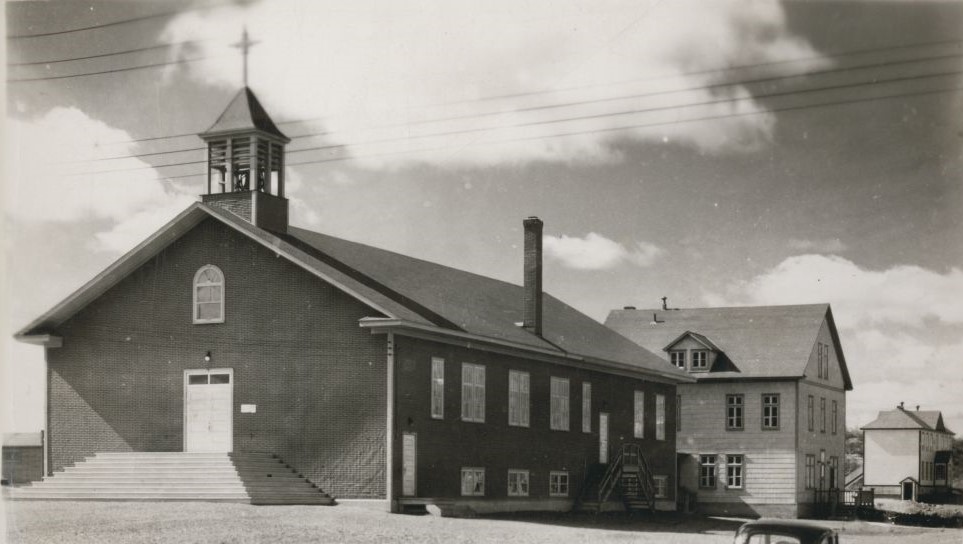 Photographie en noir et blanc d’une église recouverte de papier goudronné et surplombée d’un clocher dont la croix se confond avec les nuages. À l’arrière, un presbytère de planches de trois étages. 