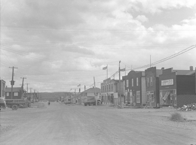 Black and white photograph of a gravel road lined with recently-constructed buildings, many with Boomtown façades. A St-Onge company truck is parked on the right.