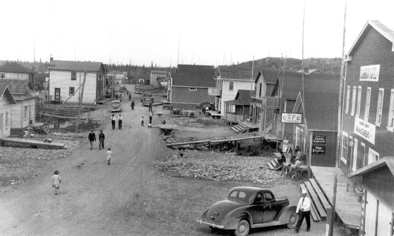 Black and white photograph of a narrow gravel road lined with rudimentary buildings. Several cars are parked and ten or so people are out and about on the public road. Long boardwalks provide access to some residences.