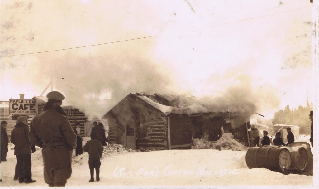 Sepia photograph of a log cabin set on fire. A large cloud of smoke fills the upper half of the photograph. A dozen curious onlookers attend the scene. On the left is a sign, 