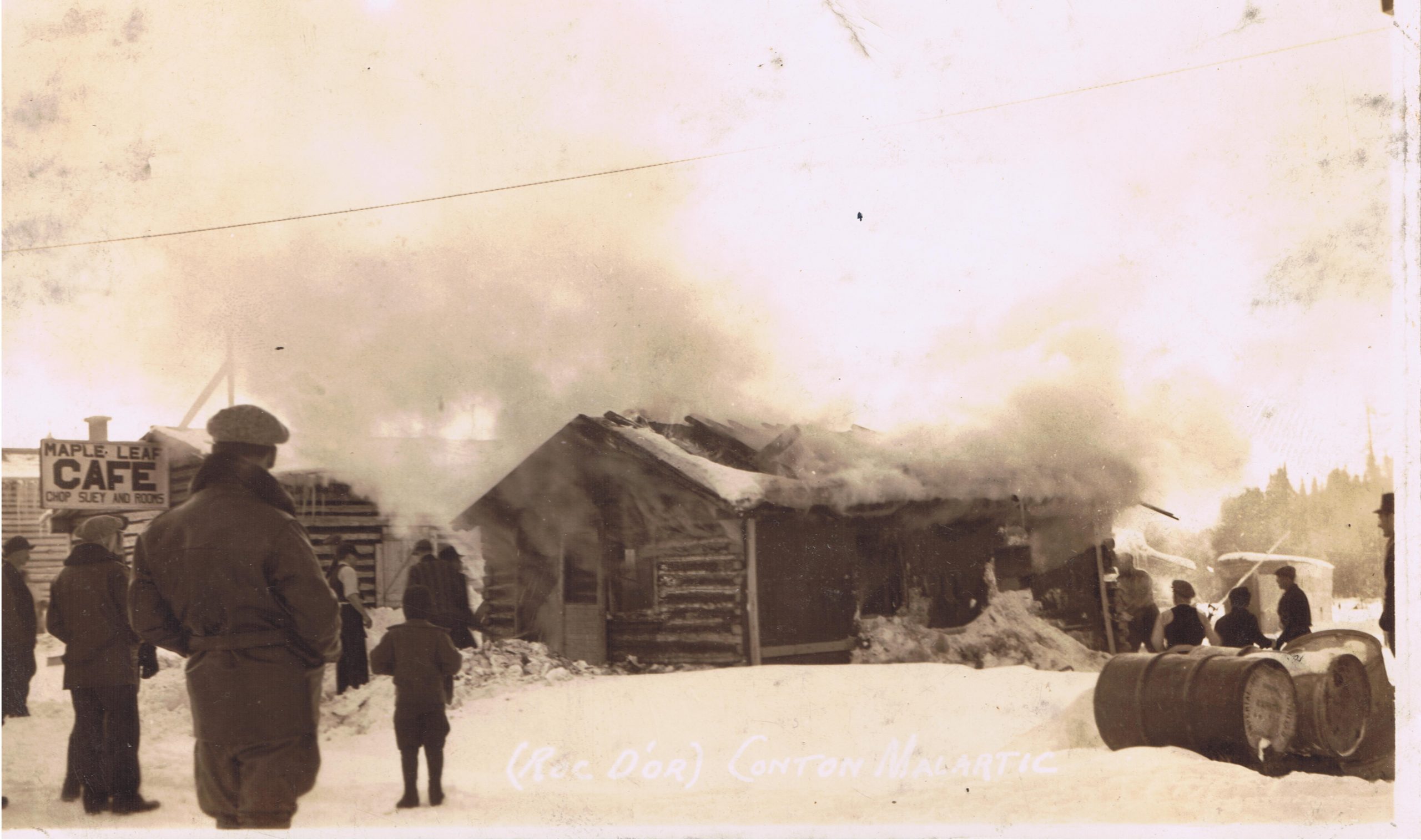 Sepia photograph of a log cabin set on fire. A large cloud of smoke fills the upper half of the photograph. A dozen curious onlookers attend the scene. On the left is a sign, "Maple Leaf CAFE Chop Suey and Rooms".