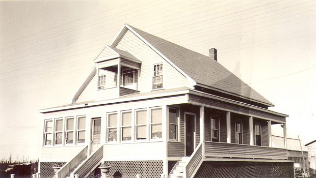 Sepia photograph of a beautiful two-storey residence. There are many windows in the front and a veranda on the right. A small balcony upstairs. In the background, a stable and a garage.