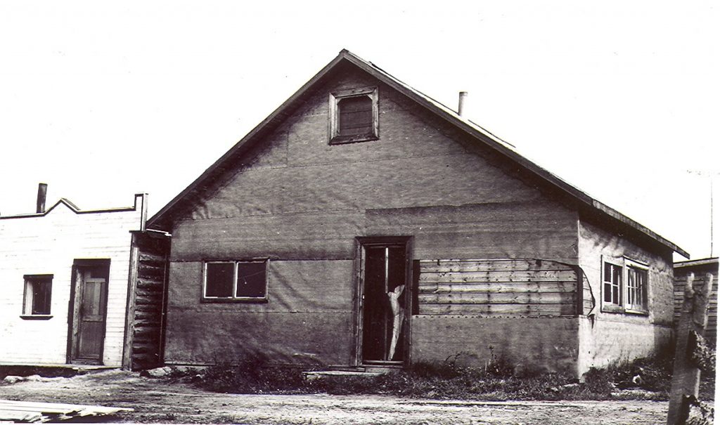 Photographie en noir et blanc d’un bâtiment recouvert de papier goudronné, dont une partie est arrachée. Quatre fenêtres, deux sur le côté droit et deux devants, sont visibles. Une cabane en bois rond avec façade de style Boomtown à gauche.