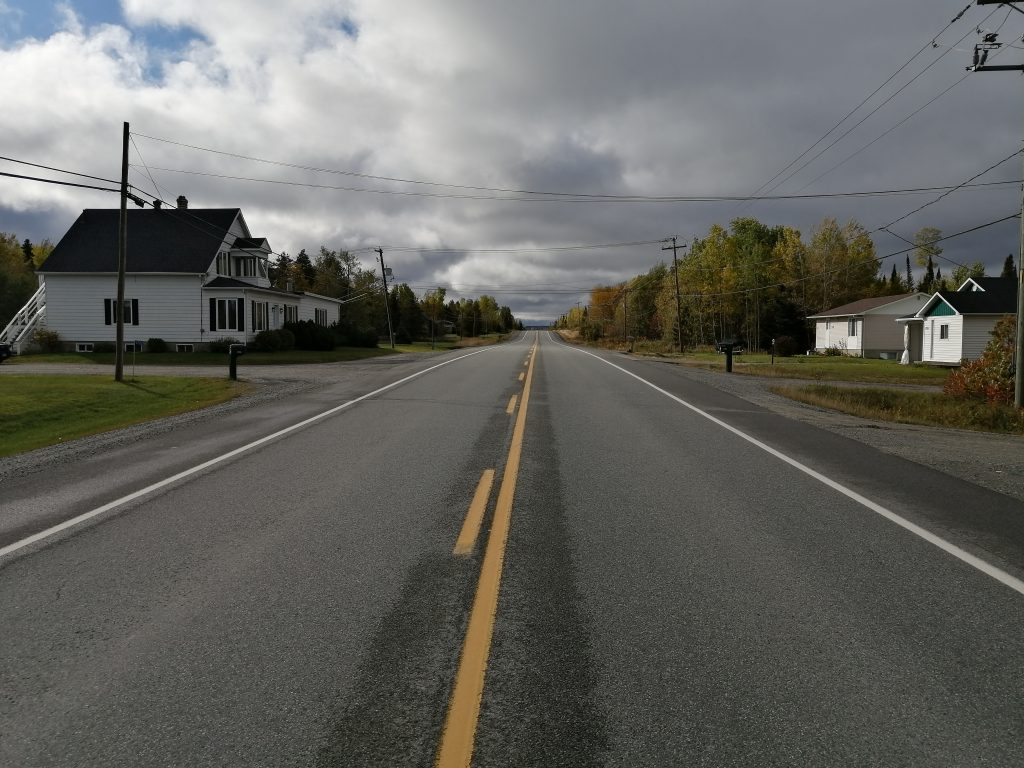 Colour photograph of an asphalt road with some residences on either side. A two-storey white house on the left and two residences on the right. The sky is cloudy.