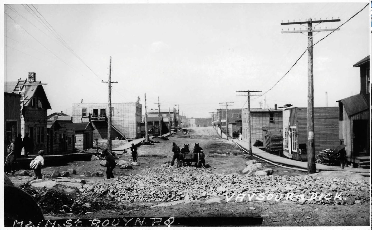 Black and white photograph of a street lined with rudimentary buildings and electricity poles. Six men and two horses work on paving. Boardwalks line the street.