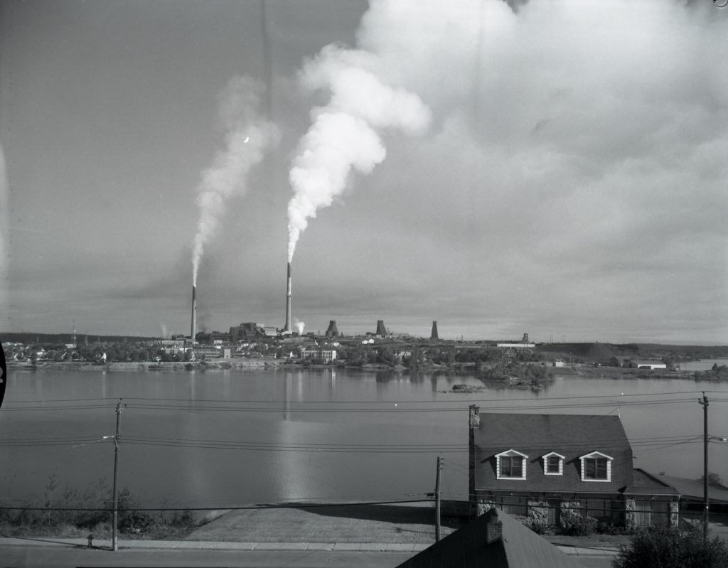 Black and white photograph of Osisko Lake with the town of Noranda, the Horne smelter, and the mine headframes in the background. In the foreground, a building used by Bergeron's Boat House.