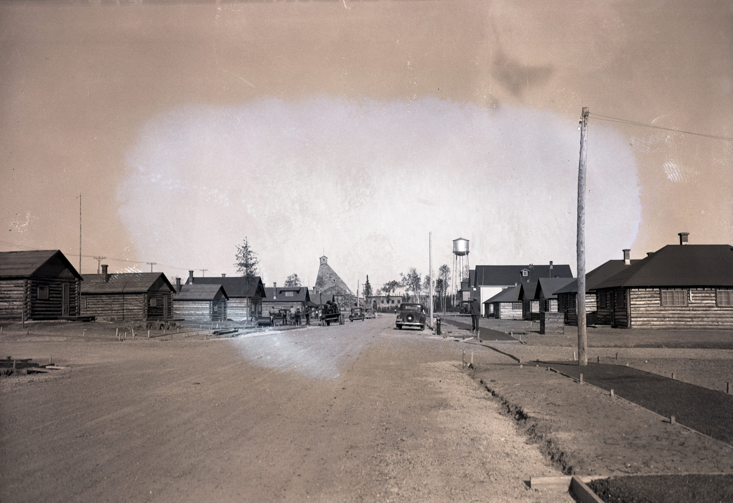 Black and white photograph of a street lined with log cabins. Men are at work on pavement. In the background, a water tower and a mine headframe.