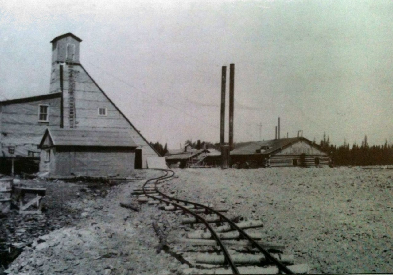 Black and white photograph of a mine headframe and a rudimentary building topped by two chimneys. In the foreground, a winding railway.
