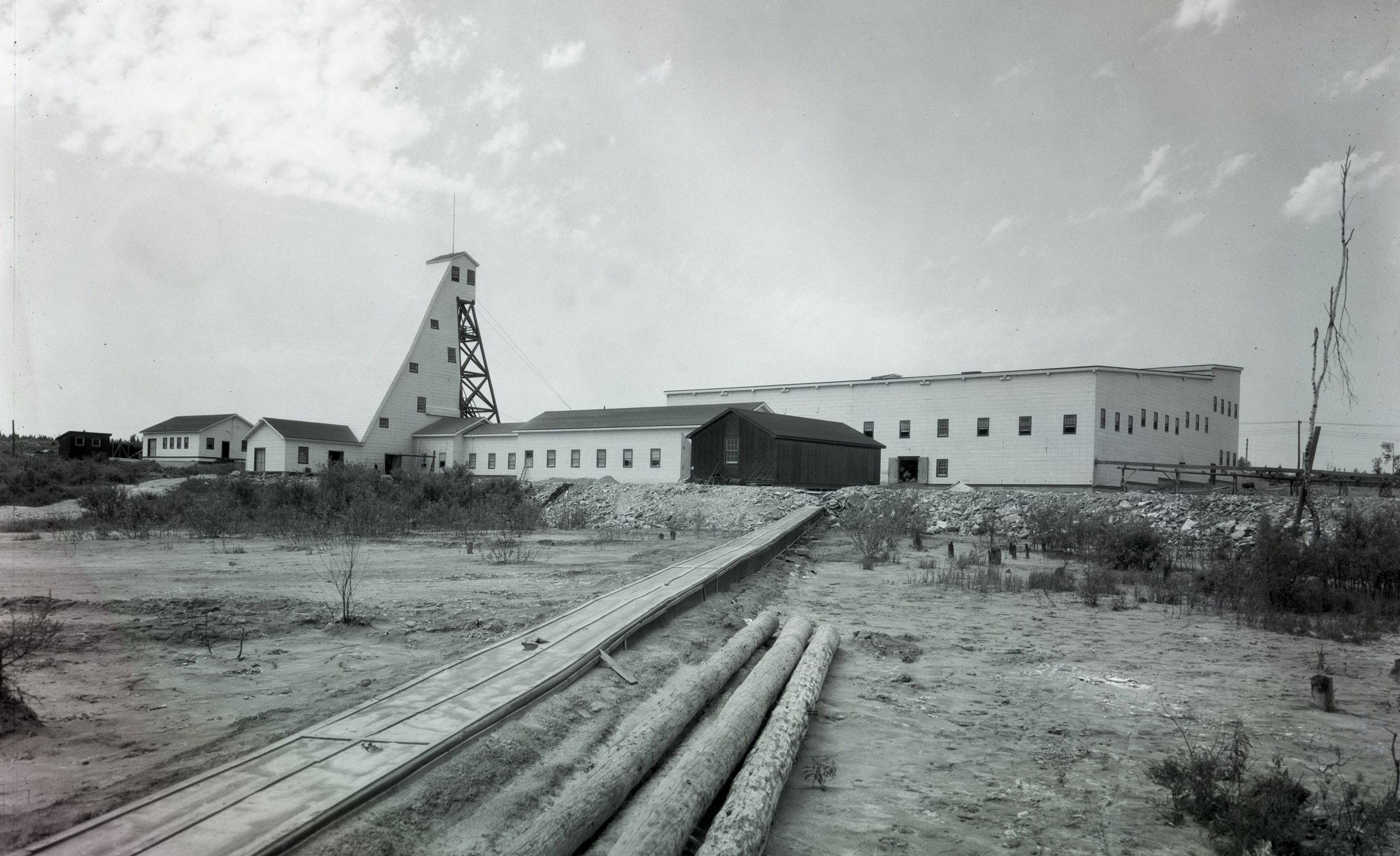 Black and white photograph of the infrastructures of Canadian Malartic Mines, including a headframe of planks. In the foreground, logs and a boardwalk provide access to the buildings.