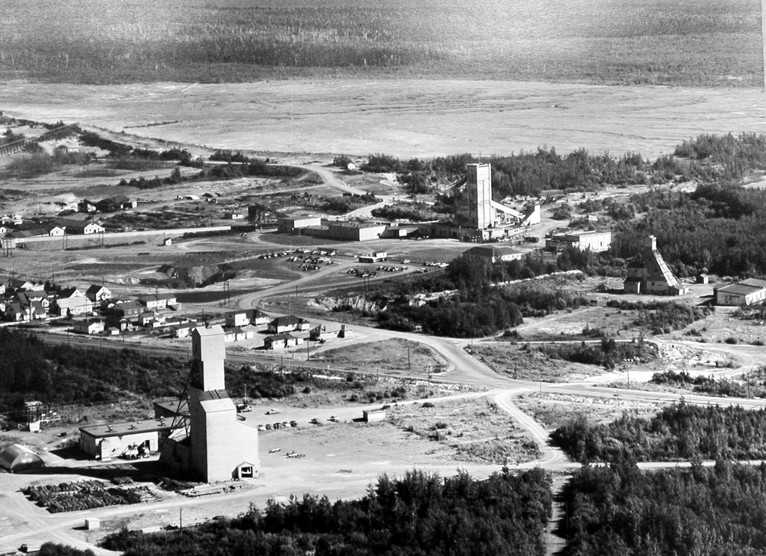 Black and white photograph of the installations of three mines, including their headframes, taken from a plane. In the centre, several houses of the private village of Halet. In the background, one of East Malartic’s mine tailings sites.