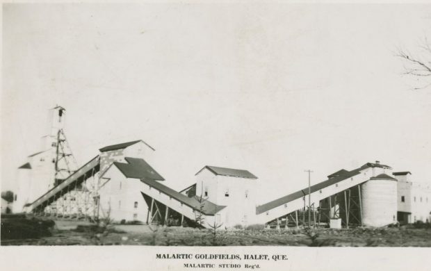 Black and white photograph of industrial buildings, including a mine headframe and several conveyors. Below, the inscription Malartic Goldfields, Halet, Que. Malartic Studio Reg'd.