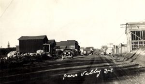 Black and white photograph of a road lined with plank buildings on which walk several pedestrians. In the foreground, a taxi cab. At the bottom, an inscription in white: 