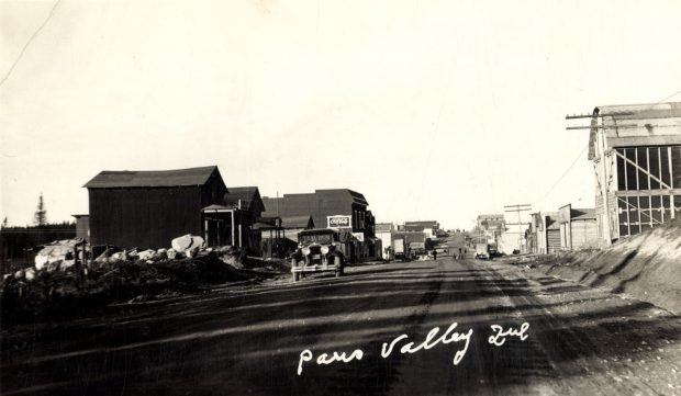 Black and white photograph of a road lined with plank buildings on which walk several pedestrians. In the foreground, a taxi cab. At the bottom, an inscription in white: Paris Valley Que.