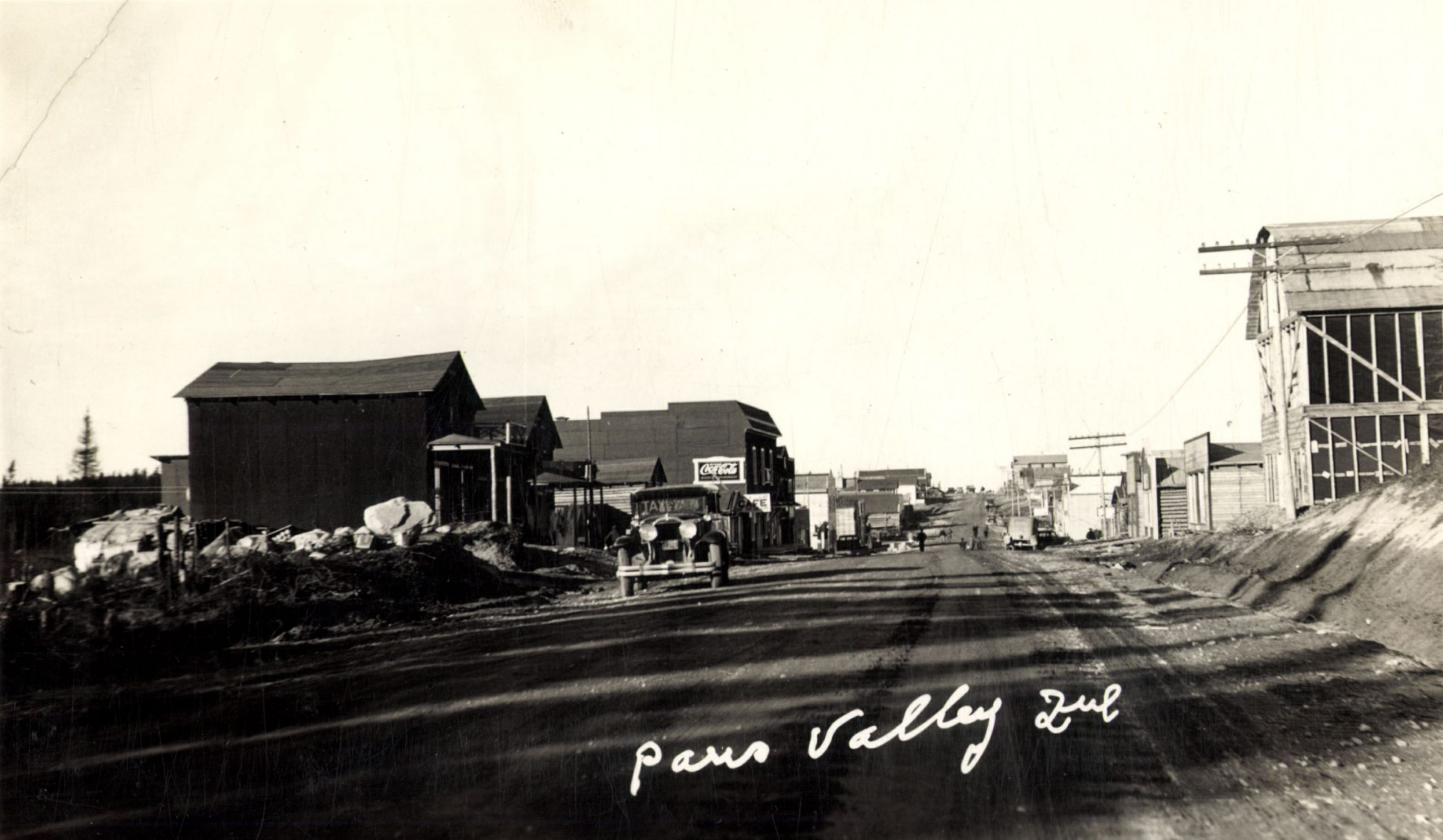 Black and white photograph of a road lined with plank buildings on which walk several pedestrians. In the foreground, a taxi cab. At the bottom, an inscription in white: "Paris Valley Que".