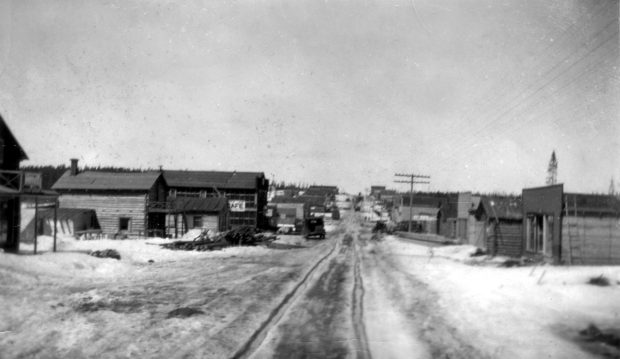 Black and white photograph of a road in winter lined with plank and log buildings. Due to the traces of traffic in the snow, the roadway is visible in the centre.