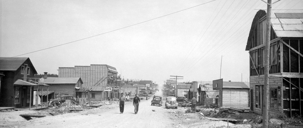 Photographie en noir et blanc d’une route de gravier bordée de bâtiments de planches ou en bois rond. Deux hommes marchent vers la caméra. À l’avant-plan, à droite, un poteau téléphonique. 