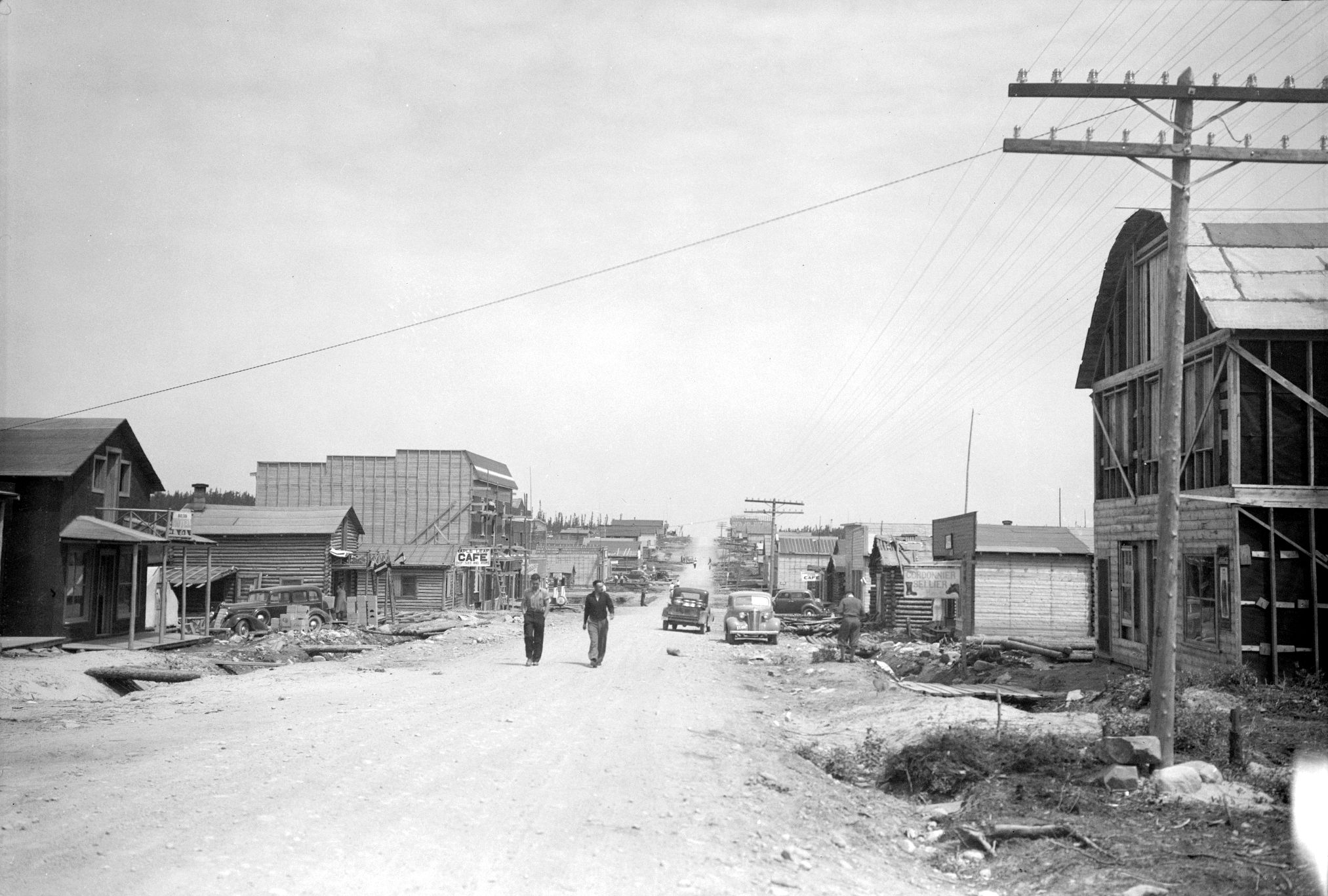 Black and white photograph of a gravel road lined with plank and log buildings. Two men walk toward the camera. In the foreground, on the right, a telephone pole.