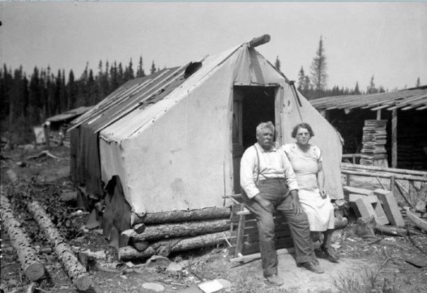 Black and white photograph of an elderly couple in front of a rudimentary dwelling: a wood and textile tent. Log cabins are visible on the right and in the background.