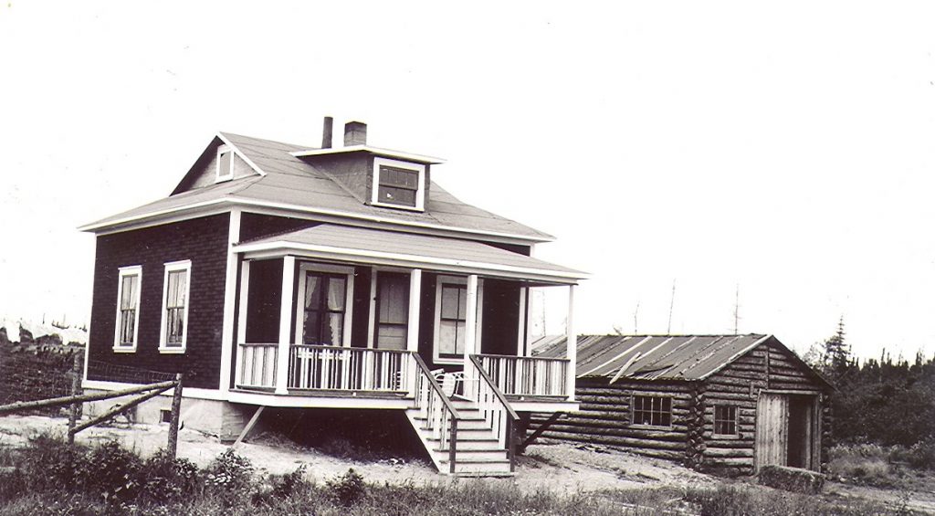 Photographie noir et blanc d’une maison propre et moderne avec galerie à l’avant. À droite, un garage rudimentaire en bois rond.