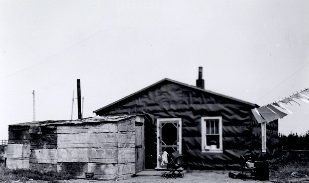 Photographie en noir et blanc d’une habitation rudimentaire divisée en deux sections : La première est recouverte de papier goudronné et la seconde de feuille de métal. Il y a une carrosse devant la maison et une corde à linge. 