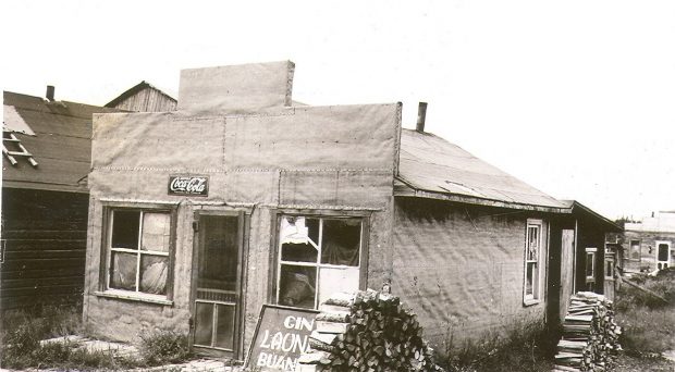 Black and white photograph of a tar paper house with a Boomtown façade. A poster placed on the floor reads, Gin LAUNDRY buanderie.