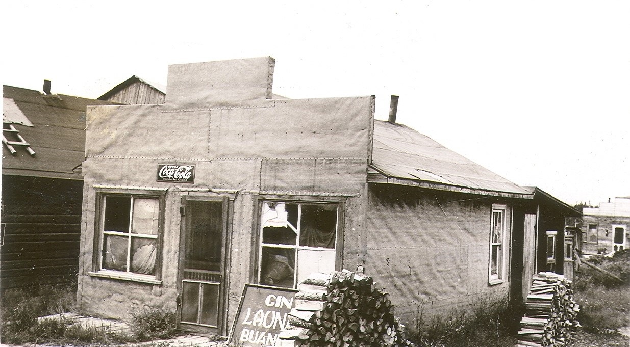 Black and white photograph of a tar paper house with a Boomtown façade. A poster placed on the floor reads, "Gin LAUNDRY buanderie".