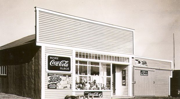 Black and white photograph of a plank house with a Boomtown façade on which several Coca-Cola advertisements are displayed. Several products are displayed in the window.