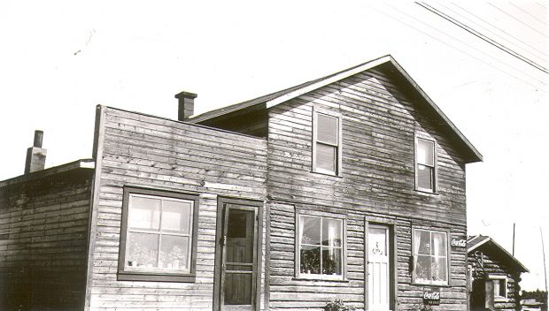 Black and white photograph of a plank building made up of two dwellings. Two Coca-Cola advertisements are displayed on the building. On the right, a log cabin.