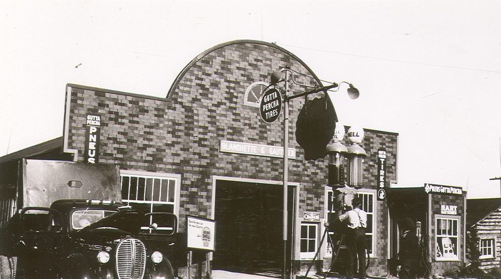Photographie en noir et blanc d’un garage dont la devanture est recouverte de papier goudronné. Au-dessus de la porte de garage, l’enseigne « Blanchette & Gauthier ». À l’avant, un homme qui actionne les pompes à essence. À gauche, une voiture avec une remorque.