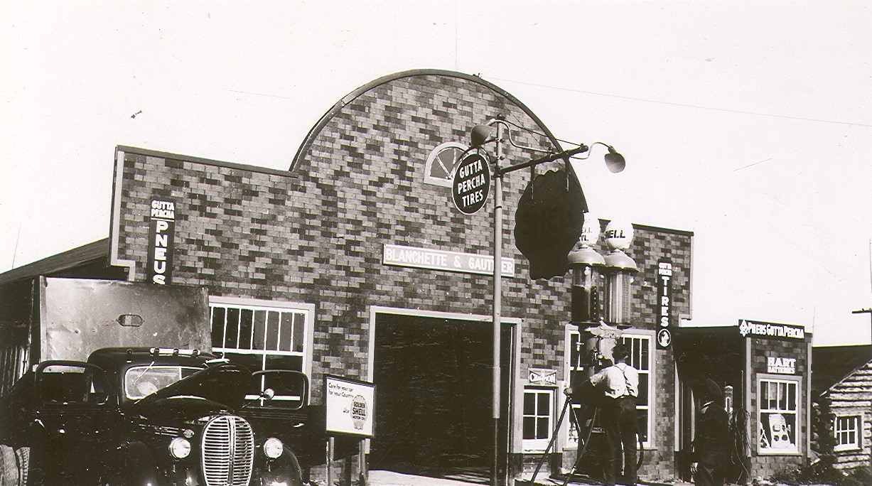 Black and white photograph of a garage with a tar paper front. Over the garage door, the "Blanchette & Gauthier" sign. In front, a man operating the gas pumps. On the left, a car with a trailer.