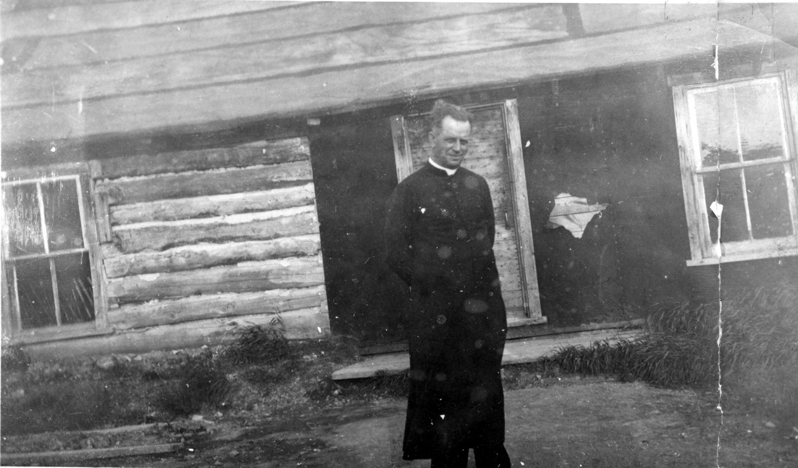 Black and white photograph of a man in a cassock posing in front of a log cabin partially covered with tar paper.