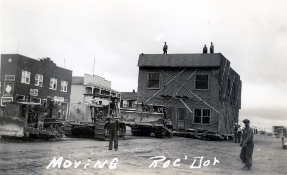 Black and white photograph of a two-storey building being moved by three tractors. Several men are on the roof. Several curious onlookers are at the scene. In the background, several buildings.