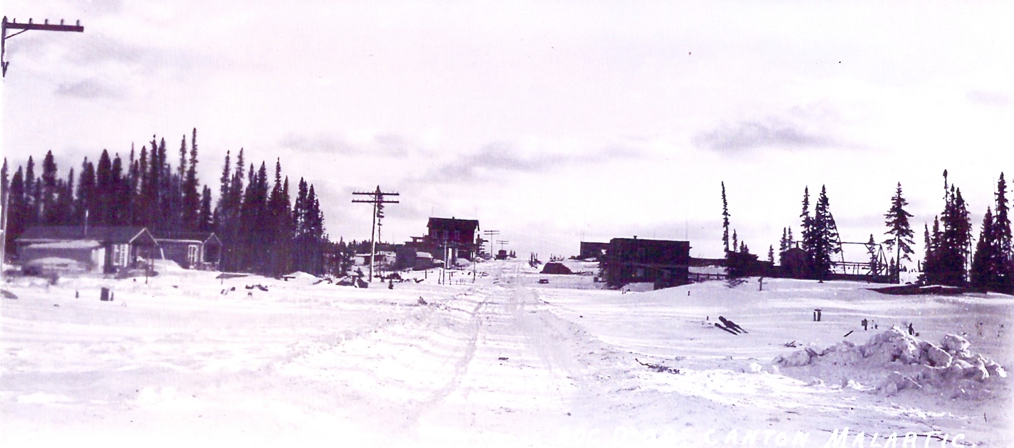 Black and white photograph of a snowy hamlet with a road down the centre. Log and plank buildings and telephone poles are visible.