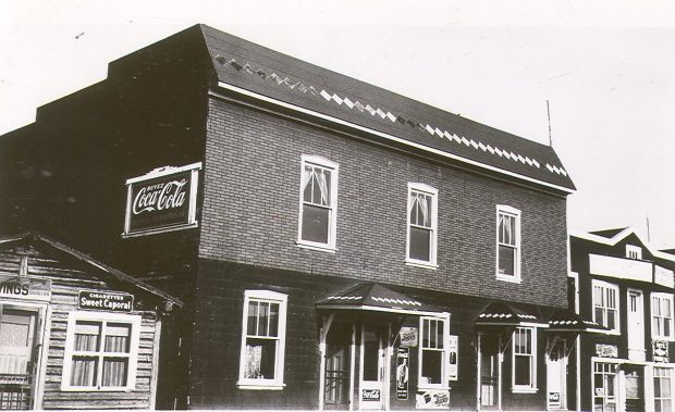 Black and white photograph of a two-storey building covered with tar paper. The front has six windows and four doors. On the side, a “Drink Coca-Cola” advertisement. On the right, the Frontenac Hotel. On the left, a log cabin.