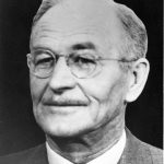 Black and white photograph of a man in a suit and tie wearing glasses and a thin moustache.